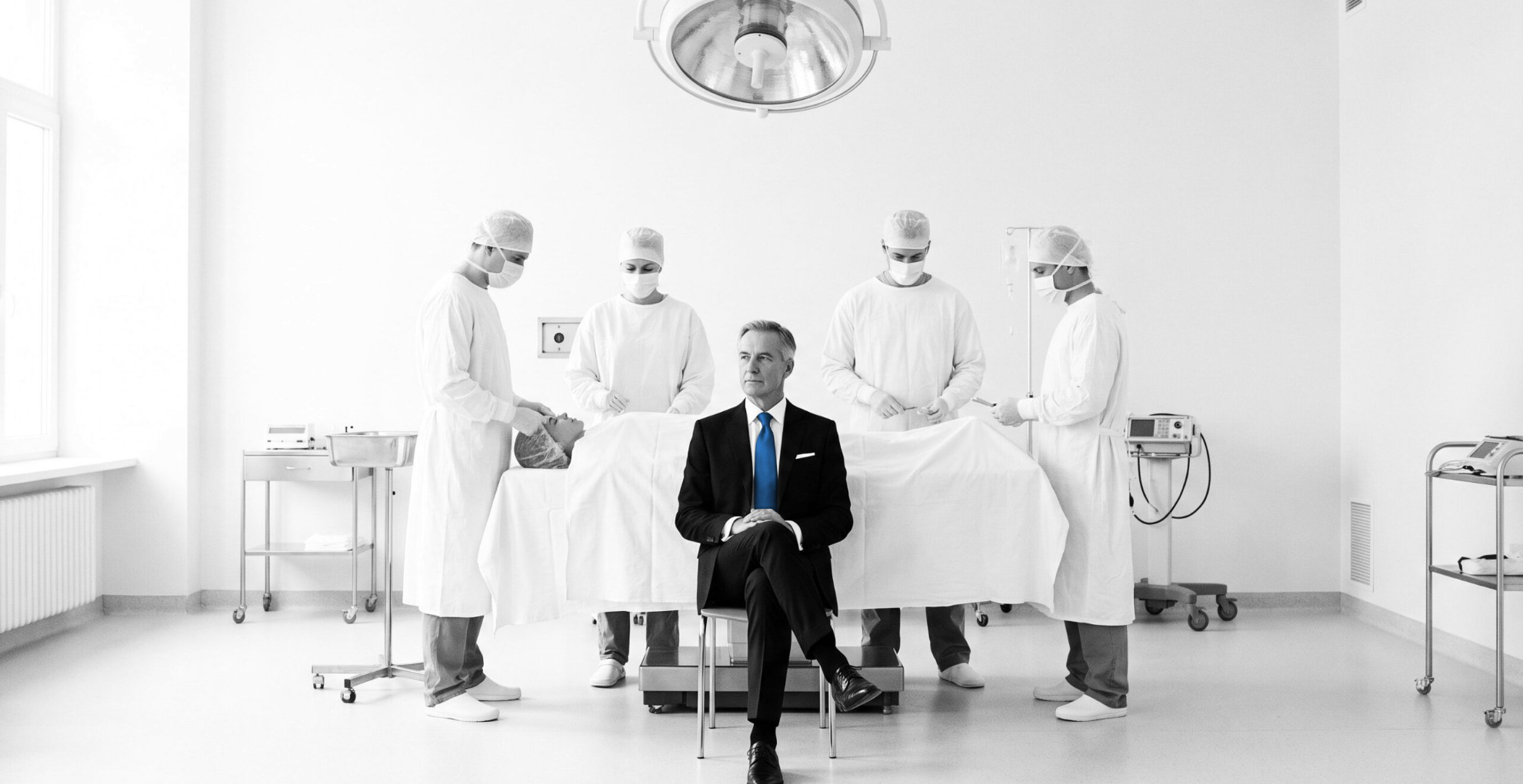 Black-and-white operating room where four surgeons work around the table and a businessman in a blue tie sits with them, the insurer inside the hospital pilot