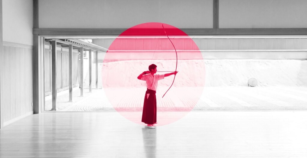 Kyudo archer at full draw in a white dojo, framed by a red circle: the aim is committed before the arrow leaves the bow