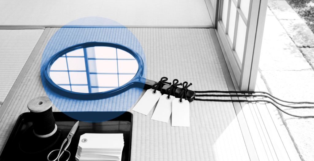 Black-and-white Japanese room: a round lacquer hand mirror lies on tatami with three blank paper tags tied to its handle, their black cords running out past a shoji into the garden, beside a tray holding the same cord and a stack of spare tags.