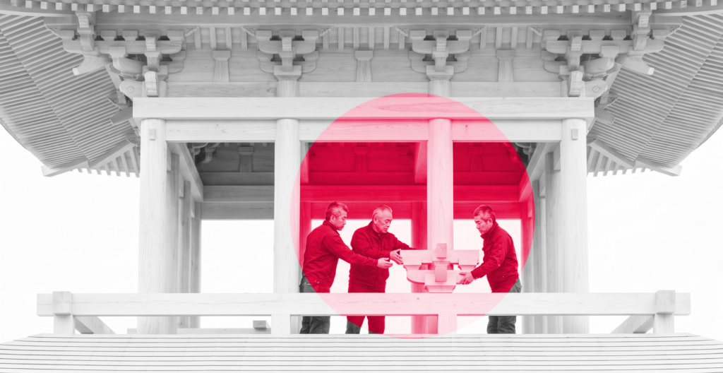 Black-and-white photograph of a wooden Japanese pagoda under construction: an older master carpenter guides two greying carpenters fitting a bracket on the bare middle storey, framed by a crimson circle.