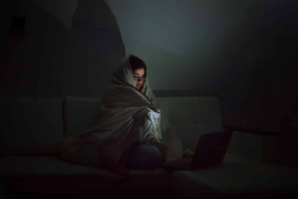 A young woman sits in a dark room looks at the laptop screen, wrapped in a blanket, burnout syndrome