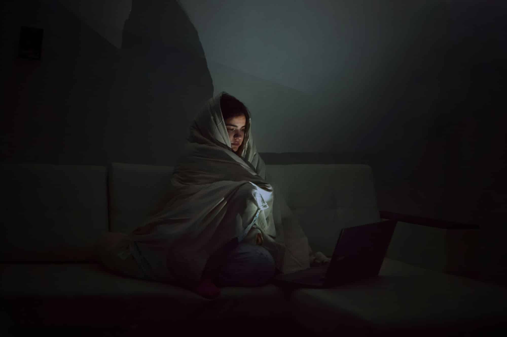 A young woman sits in a dark room looks at the laptop screen, wrapped in a blanket, burnout syndrome