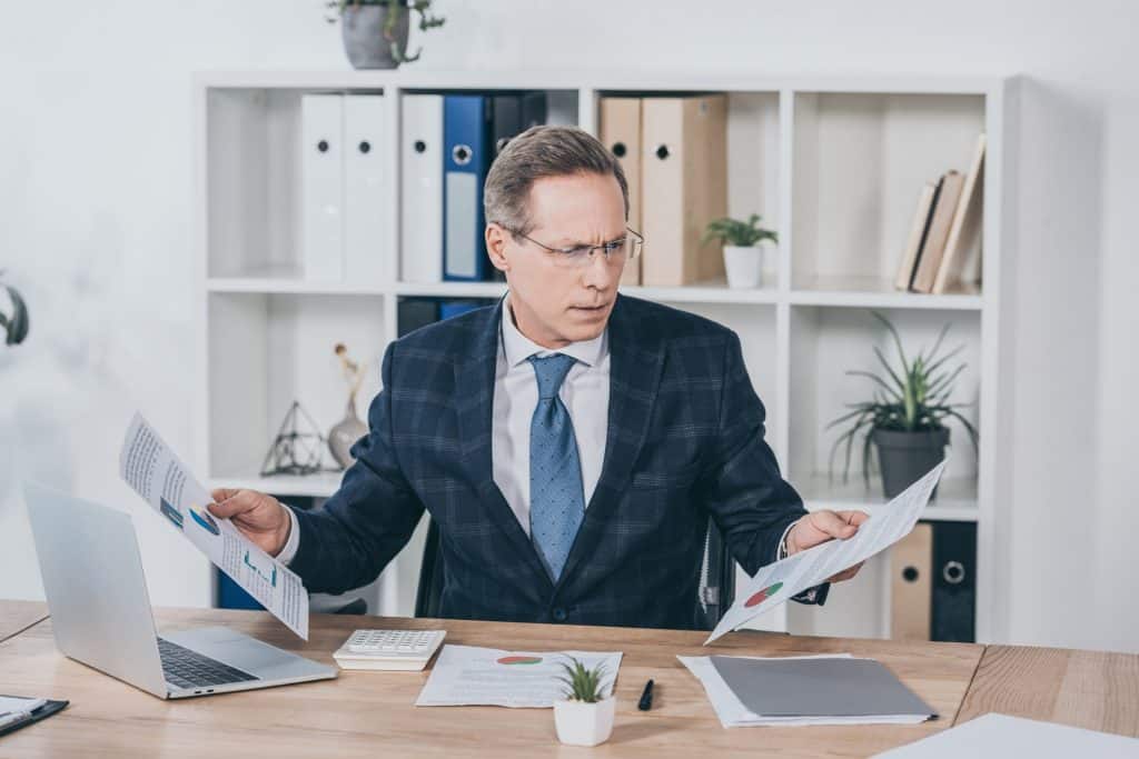 middle aged businessman sitting at table, holding documents and reading in office, compensation